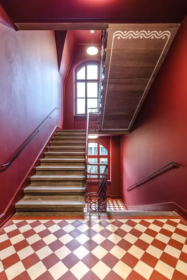 Vue dans la cage d’escalier entièrement recouverte de rouge. En sous-face, les volées d’escalier sont agrémentées d’ornements discrets de couleur blanche. Un sol  en dalles posées en losange sur les paliers.
