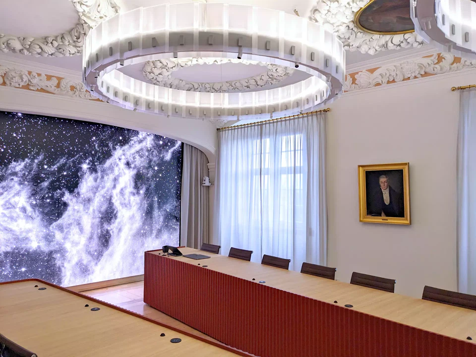 Die beiden Kameras und Lautsprecher links und rechts am Bogenpilaster und die grosse LED-Wall sind Teil der neuen Konferenzinfrastruktur. Tischmikrofone und Steckdosen sind in die Tischfläche integriert.