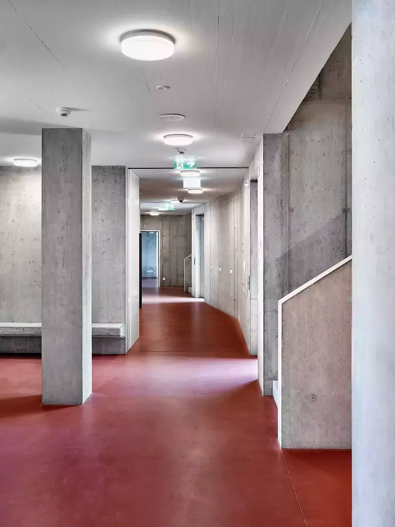 Blick in den Eingangsbereich des Wohntrakts mit roh geschaltem Beton und rot abgesetztem Bodenbelag.