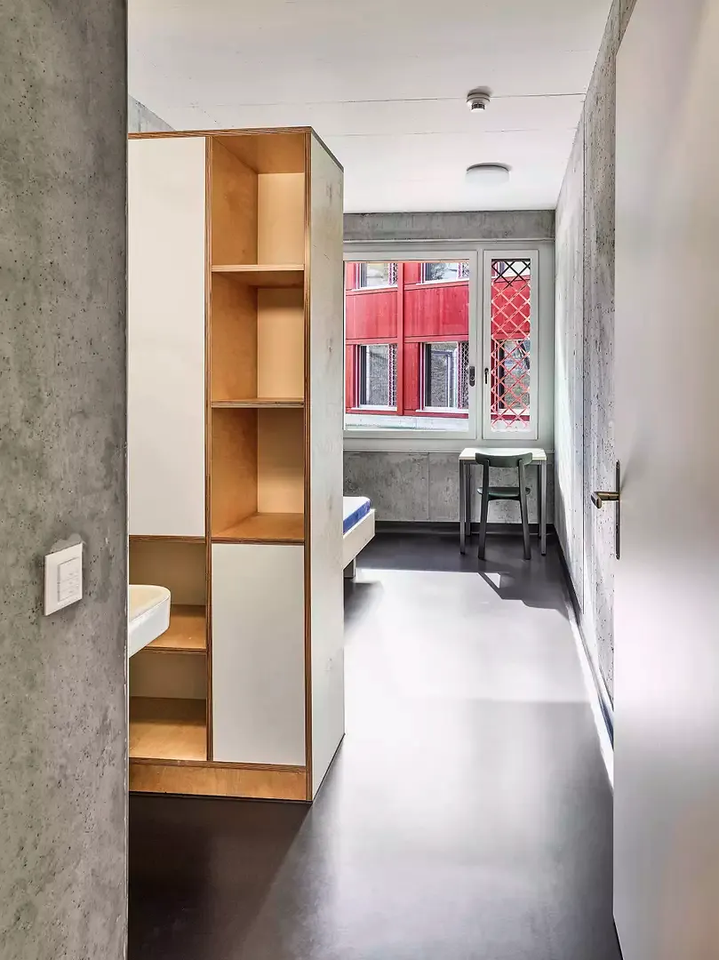 Blick in ein Einzelzimmer  des Wohntrakts, das zweckmässig und solide ausgestattet ist mit Waschgelegenheit,  Regal- und Schrankwand.  Auf der Rückseite geschützt das Bett kombiniert mit einem Sitzplatz am Fenster.