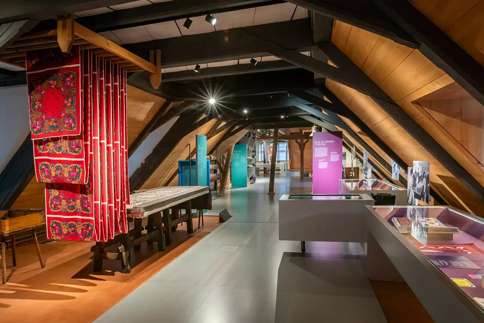 Des vitrines argentées et des éléments encastrés sur des planches qui semblent flotter s'étendent en escalier dans toute la salle d'exposition et renforcent la luminosité des textiles aux couleurs vives.