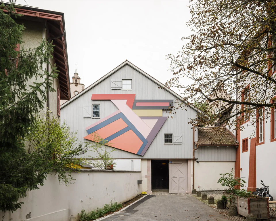 Museumsplatz und Westfassade des Jüdischen Museums Schweiz in Basel mit Jeziory, dem Werk des US-amerikanischen Künstlers, Frank Stella. Die Wand des Platzes ist mit mittelalterlichen Grabsteinen der Dauerausstellung Kult, Kultur, Kunst bespielt.