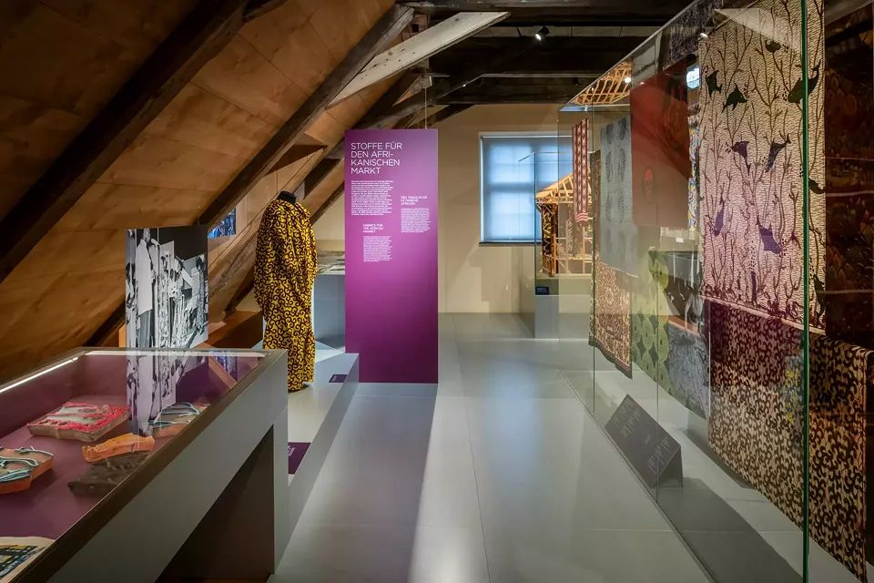 Dans la vitrine de table à gauche sont présentés des modèles en bois pour l'impression de tissus destinés au marché africain, dans la vitrine en verre et drapés sur le mannequin de couture les tissus imprimés. 