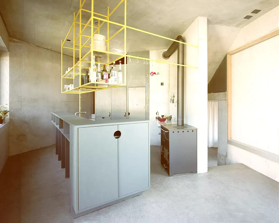 Die Küche ist in die Promenade architecturale eingewoben. Ein holz­gefeuerter Ofen dient zum Kochen und Heizen. Das hängende Metallgerüst  ist Regal, Beleuchtung  und Dampfabzug in einem  und zoniert zugleich  subtil den Kochbereich.