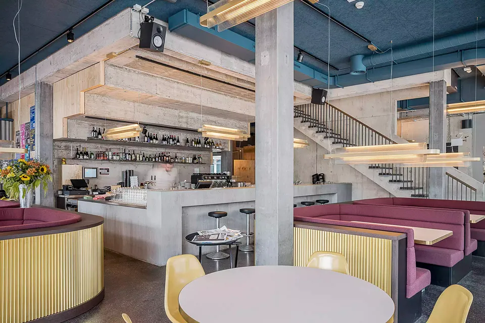 Bistro/bar: vue de la zone bistro au rez-de-chaussée, le bar entouré de béton brut est  situé sous les grands escaliers, les tables individuelles et les niches avec des banquettes rembourrées assurent détente et convivialité.