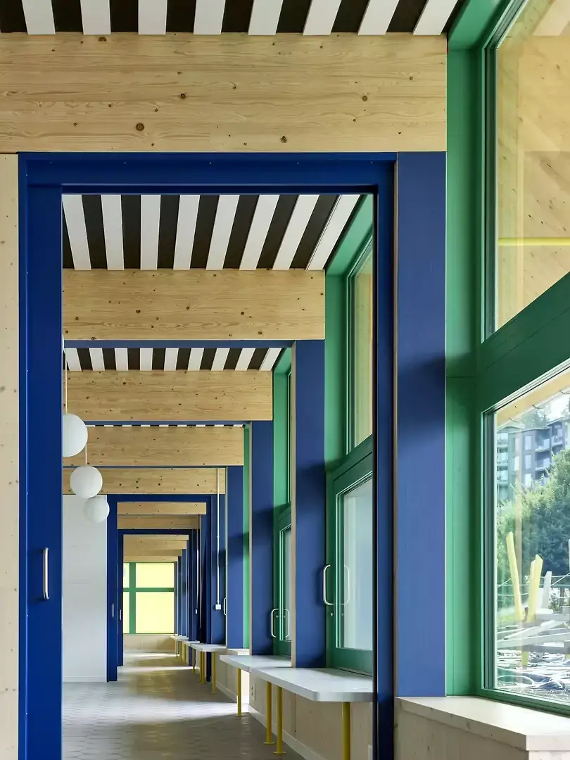 Fenêtres vertes, portes  bleues combinées avec des éléments porteurs en bois  et des surfaces blanches:  la palette chromatique marquante des logements  se retrouve dans l’école enfantine.