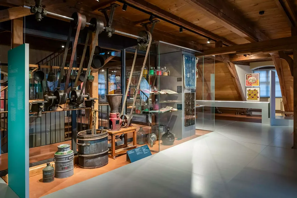 Grandes vitrines en verre avec divers outils et ustensiles pour la gravure de modèles ainsi que des bocaux contenant des pigments et des livres d'échantillons de couleurs. À côté, un moulin à peinture, une cuve de mélange de peinture et l'engrenage de tra