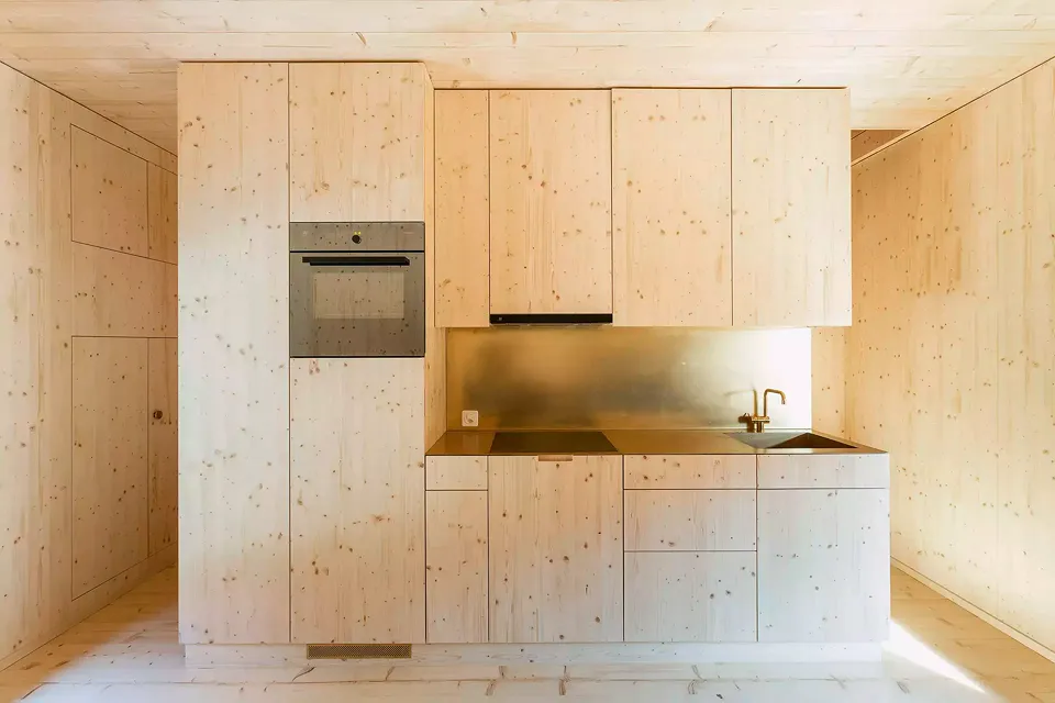 Le revêtement de la niche de cuisine, le mélangeur et l’évier sont réalisés en laiton, un matériau brut qui se patine et porte ainsi la marque du temps, en analogie au vieillissement naturel du bois.