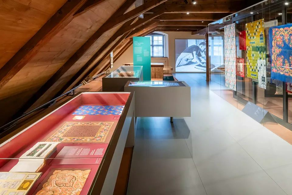 Vitrine de table avec des livres d'échantillons et des exemples de foulards. A droite, une vitrine avec de grands échantillons de tissus et des foulards avec des sujets illustratifs. En arrière-plan, la projection sur tout un mur des processus artisanaux.