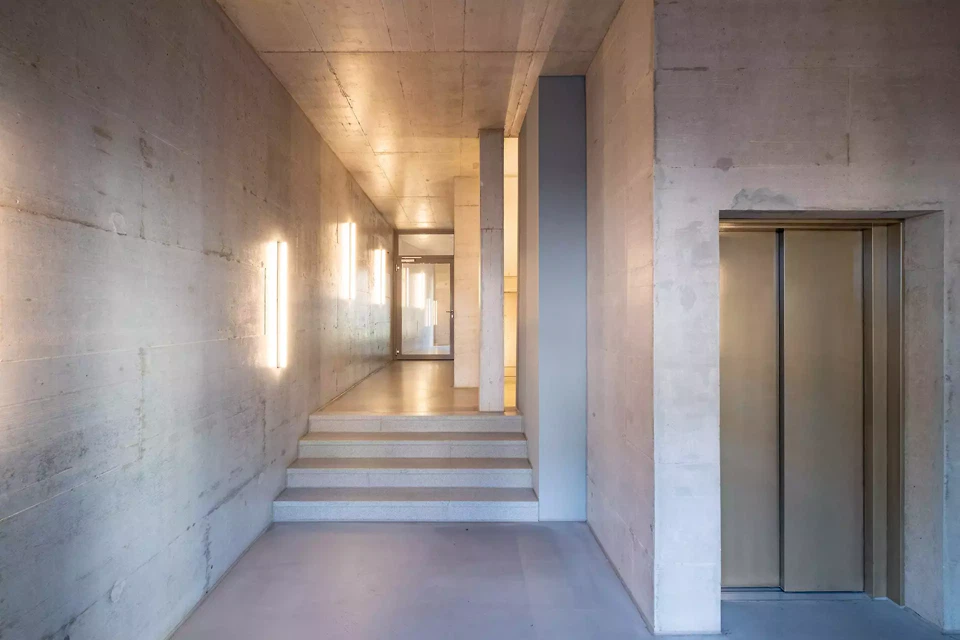 Den Eingangsbereichen und Treppenhäusern wurde bewusst Raum gelassen und ihre Gestaltung steht im Spannungsfeld zwischen der Rauheit des ruppig verarbeiteten Betons, kombiniert mit einer silbrigen Lasur und feinen Geländerdetails.