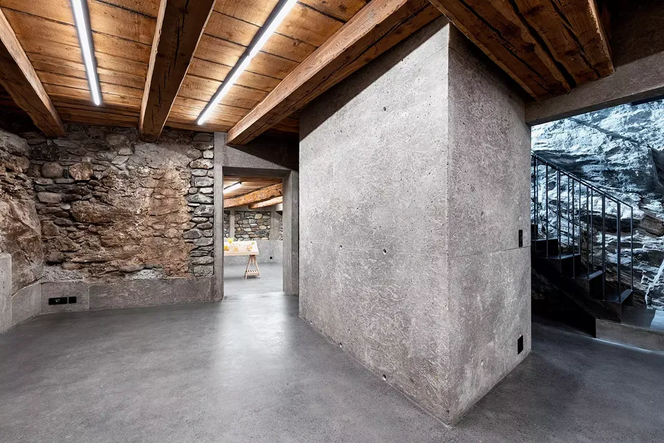 Chesa Della Santa, sous-sol: reprise en sous-oeuvre des murs en maçonnerie traditionnelle, noyau sanitaire en béton lavé, sous-face du plancher d’origine de l’étable en bois d’arve et escalier en acier longeant la paroi rocheuse existante.