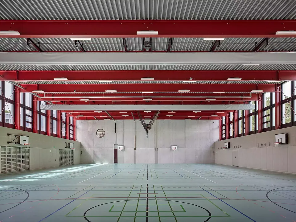 Die neue Dreifachturnhalle wird von ihren roten Stahlträgern und einem bemerkenswerten, zweiseitigen Tageslichteinfall charakterisiert.