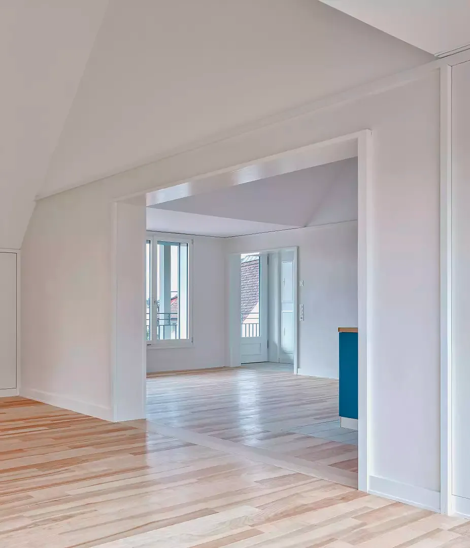 Grand appartement sous combles de 3 1/2 pièces dans le nouveau bâtiment: vue depuis la salle de séjour sur la grande cuisine ouverte (avec bloc-cuisine bleu) dont  le sol est recouvert  d’un parquet huilé en  bois de frêne.