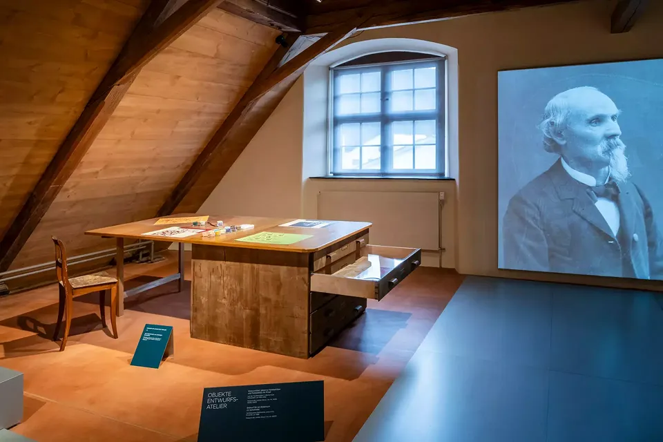 Table de travail de l'atelier de conception. Le tiroir ouvert montre un dessin original. À côté, une projection montre des images du design des tissus, de leur production jusqu'à leur présentation sur les podiums internationaux.