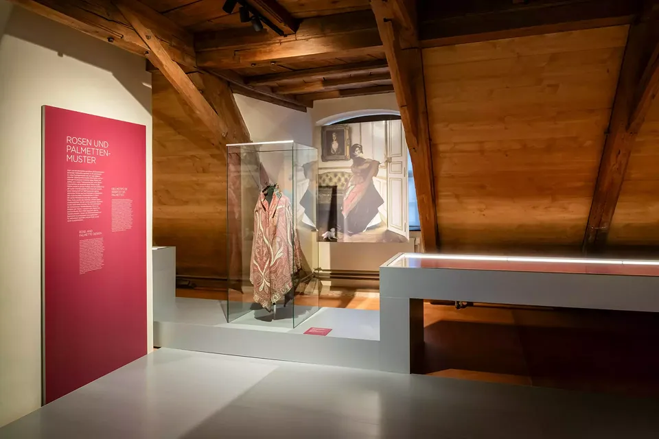 Panneau d'introduction sur le thème « Motifs de roses et de palmettes ». Dans la vitrine, une cape en cachemire est présentée, derrière laquelle est suspendue une reproduction de la même écharpe telle qu'elle était portée à l'époque. A droite, la vitrine 