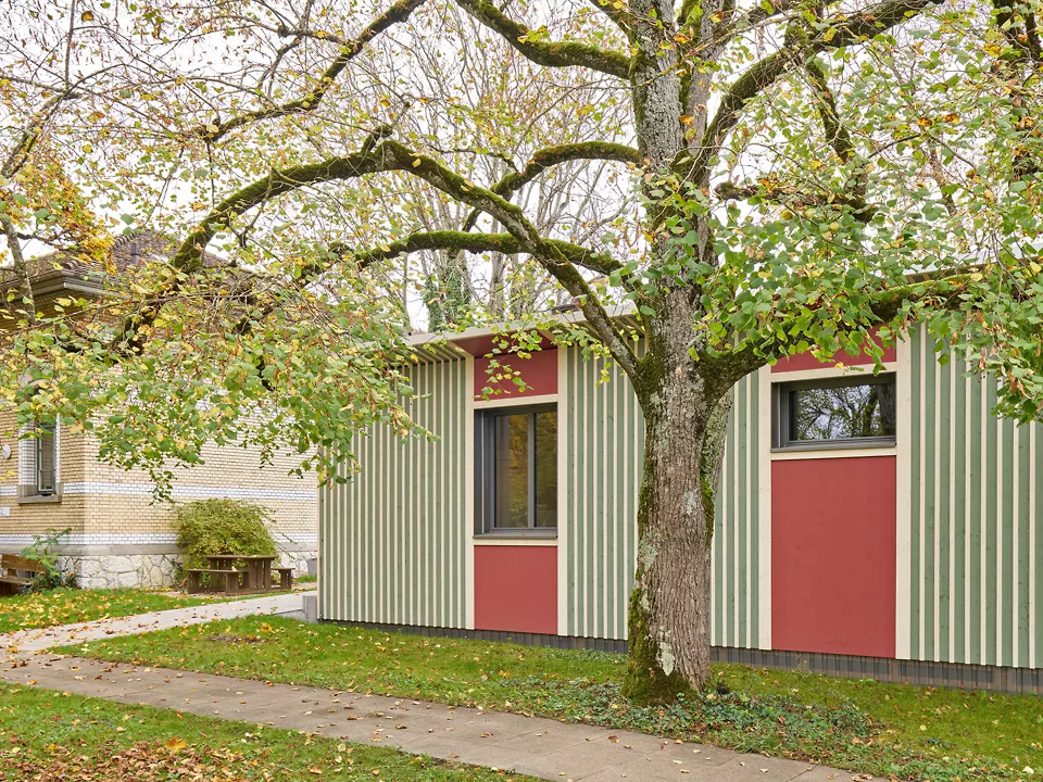 Die vertikale Holzschalung mit Deckleisten greift Material und Farbgebung der Nachbargebäude auf und interpretiert sie neu, sodass sich das Gebäude als natürliche Erweiterung in die bestehende Architektur einfügt. – Blickrichtung «Tilia Atelier Miracolo».