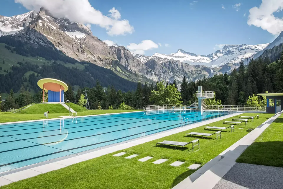 Wiederhergestelltes 50-Meter Schwimmbecken mit neuem Sprungturm und rekonstruiertem Musikpavillon vor Bergpanorama