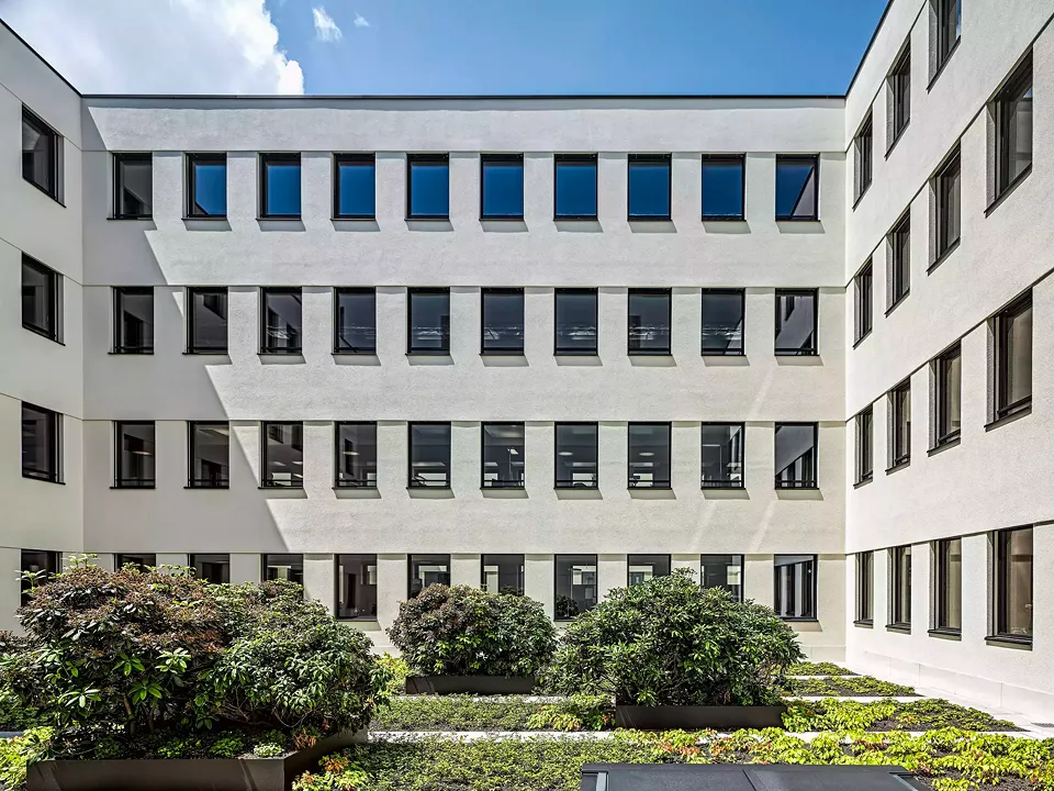 Depuis les bureaux de l’administration, la vue donne sur une vaste cour verdoyante où se trouve  une façade crépie perforée qui reflète la lumière du jour.