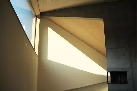 Vue depuis le salon. Le soleil du soir rentre profondément dans le bâtiment. La cheminée est encastrée dans la tête du mur porteur en béton.
