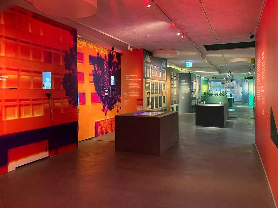Die Ausstellung startet im «Hitzeraum». Von hier öffnet sich der Blick in die Häuserschlucht der Kulissenstadt. Der erste Teil der Ausstellung ist in orangerotes Licht getaucht, diese Farbstimmung ändert sich mit jedem Abschnitt und Thema des Raums. 