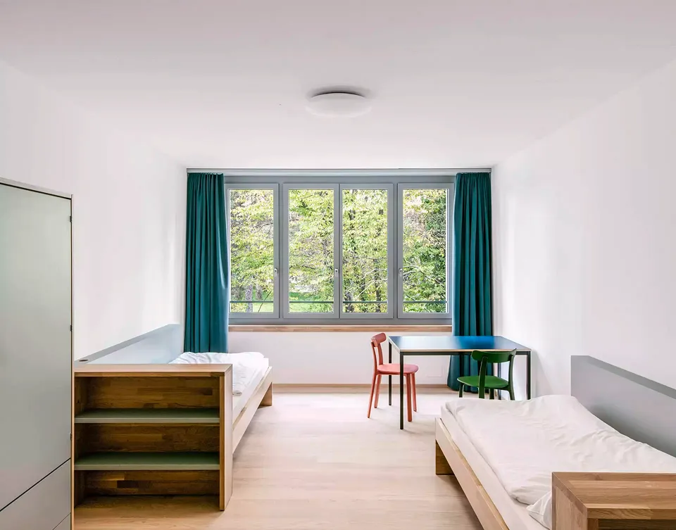 Blick in ein solide ausgestattetes Zimmer  der Wohnstation für  Kinder und Jugendliche,  die insgesamt siebzehn Zweierzimmer umfasst. Sämtliche Fensterrahmen  sind aussen dunkelgrün  und innen in einem beruhigenden und hellen Graugrün gestrichen.