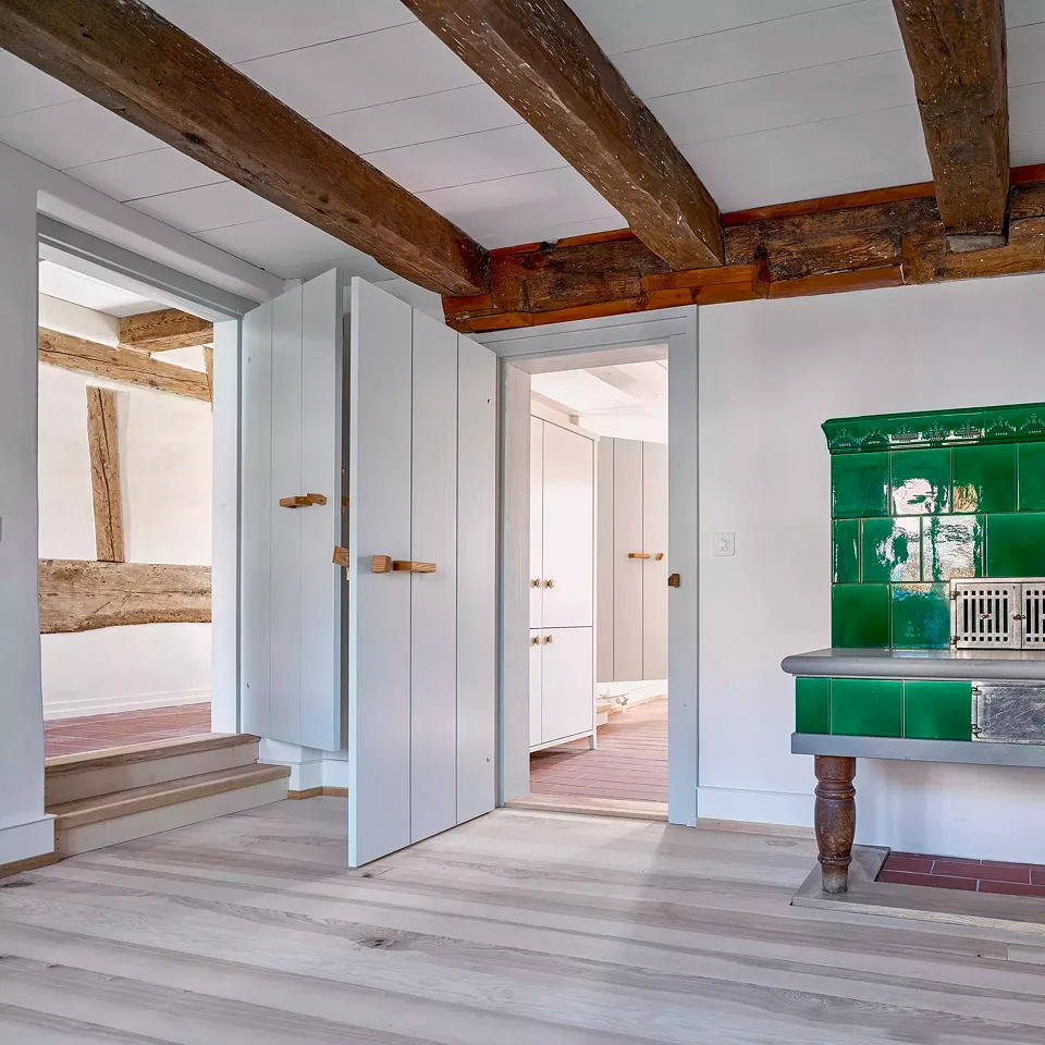 Salon de la Taunerhaus avec un vieux poêle en faïence et des portes intérieures inspirées des modèles originaux. Vue sur la cuisine avec placard et le couloir (à gauche).